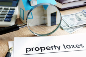 A white piece of paper that reads “property taxes” next to a magnifying glass and US $100 bills.